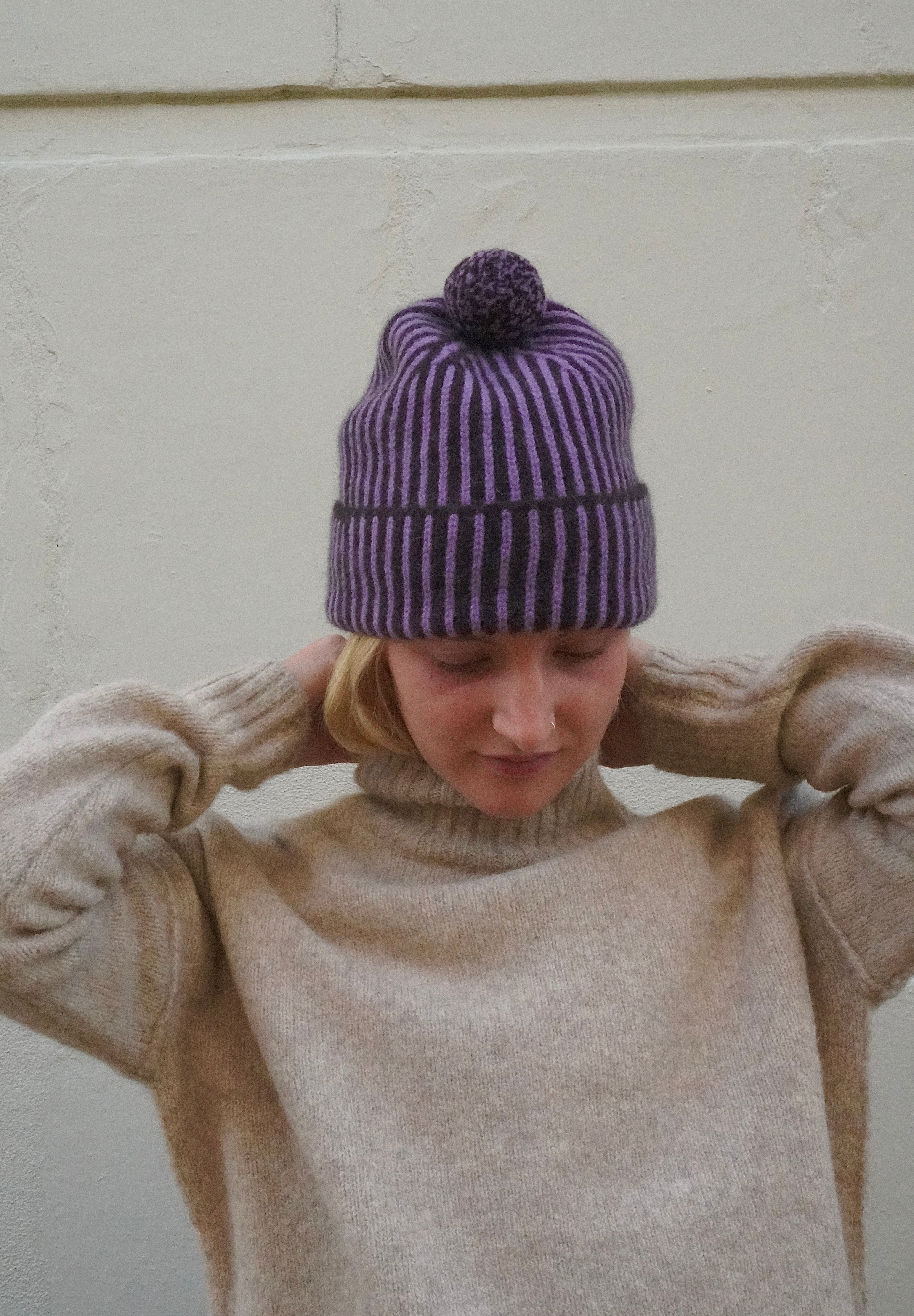 Woman wearing striped purple beanie with bobble on top, and a knitted cream polar neck jumper. 