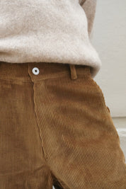 Close-up of a person wearing brown corduroy trousers and a light beige knit top against a plain off-white wall.