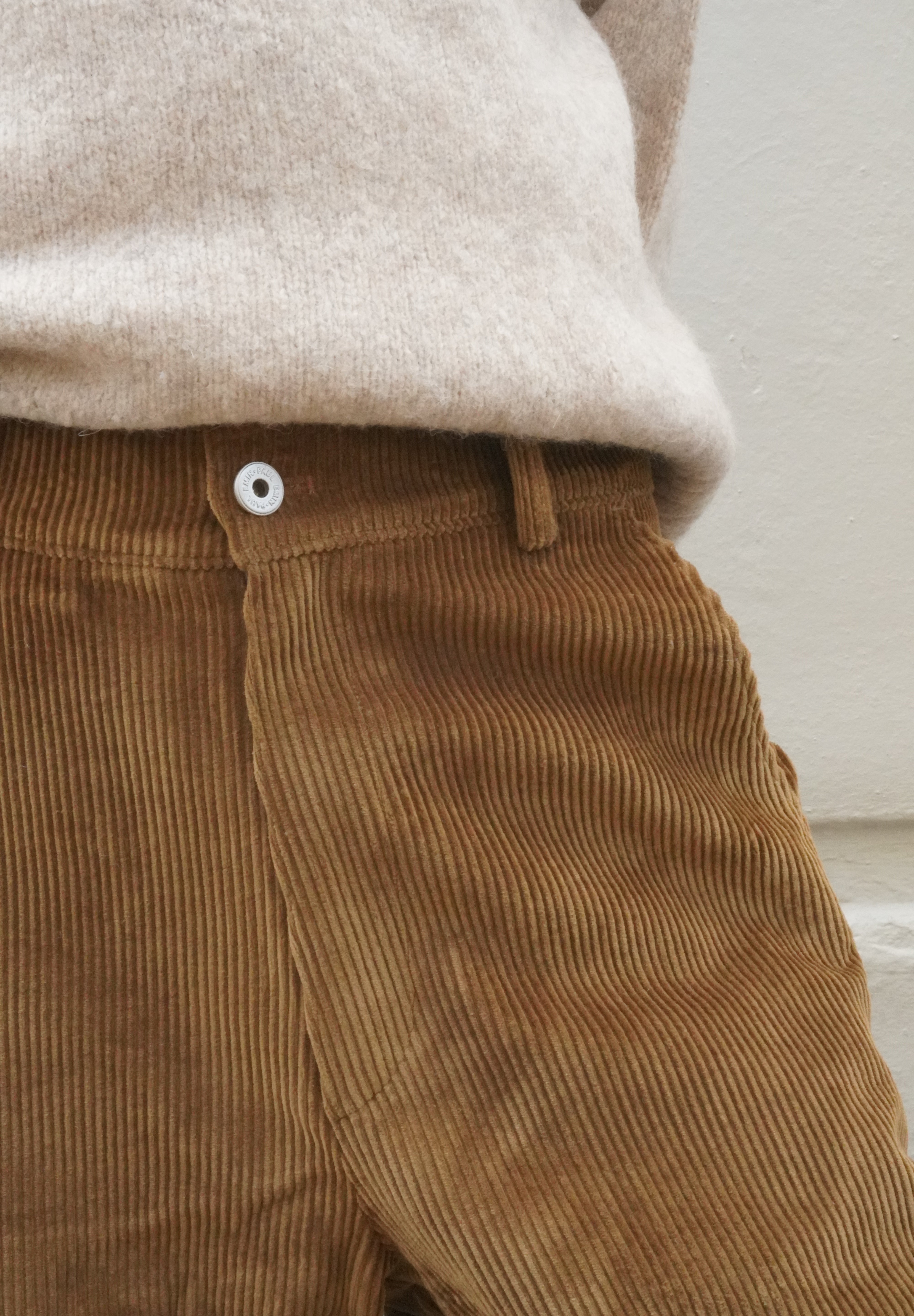 Close-up of a person wearing brown corduroy trousers and a light beige knit top against a plain off-white wall.