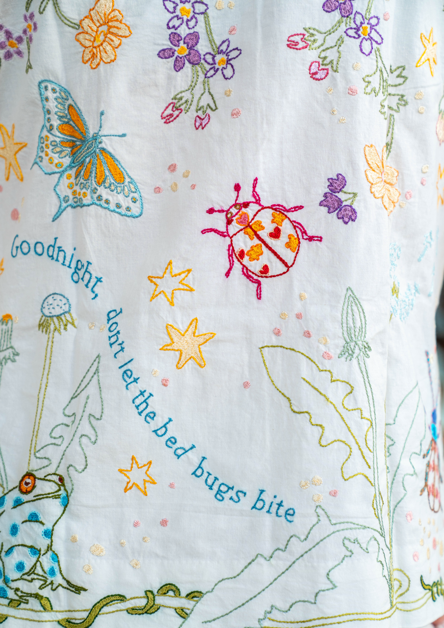 Close-up of white pyjamas with colorful embroidered wildflowers, a blue butterfly, a pink ladybug, yellow stars, a green frog, and the phrase 'goodnight, don't let the bed bugs bite' stitched in blue."goodnight, dont let the bed bugs bite"