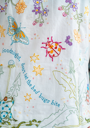 Close-up of white pyjamas with colorful embroidered wildflowers, a blue butterfly, a pink ladybug, yellow stars, a green frog, and the phrase 'goodnight, don't let the bed bugs bite' stitched in blue."goodnight, dont let the bed bugs bite"