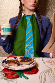 A person wearing a bright green napkin around their neck styled like a blue and yellow striped tie, sitting at a table with a plate of toast, a mug, and a newspaper against a glittery gold background.