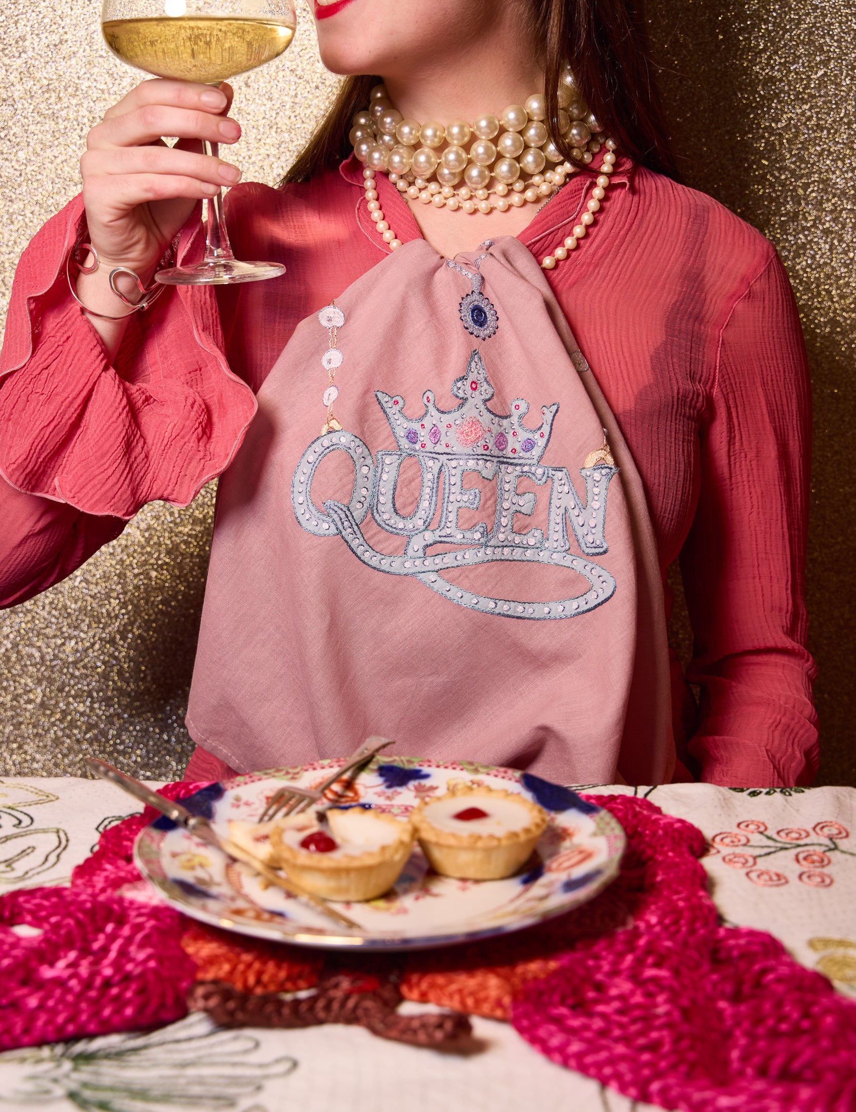 A woman in a pink top and pearl necklaces holds a glass of white wine while wearing a dusty pink napkin around her neck with 'QUEEN' and a crown design in rhinestones. A plate of mini tarts sits on a patterned tablecloth in front of her against a glittery gold background. All products from The Jacksons