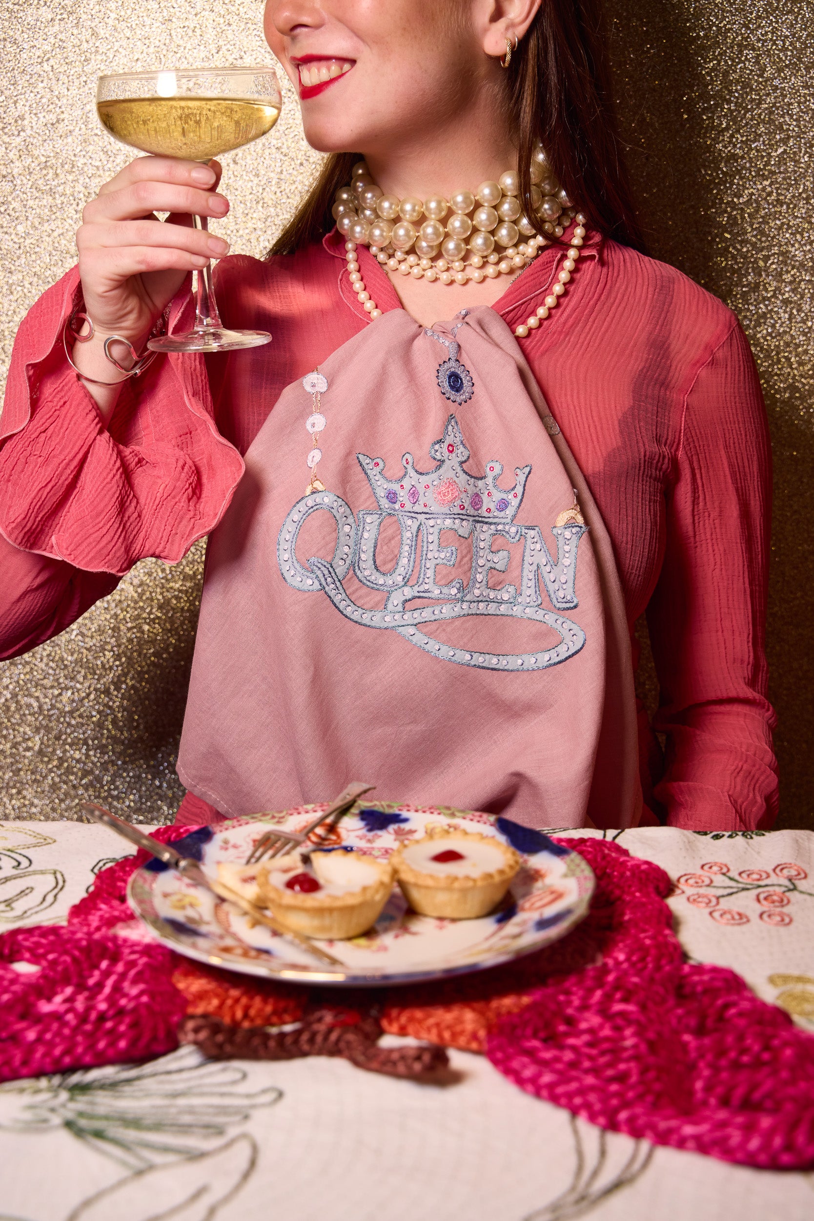 A woman in a pink top and pearl necklaces holds a glass of white wine while wearing a dusty pink napkin around her neck with 'QUEEN' and a crown design in rhinestones. A plate of mini tarts sits on a patterned tablecloth in front of her against a glittery gold background. All products from The Jacksons
