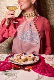 A woman in a pink top and pearl necklaces holds a glass of white wine while wearing a dusty pink napkin around her neck with 'QUEEN' and a crown design in rhinestones. A plate of mini tarts sits on a patterned tablecloth in front of her against a glittery gold background. All products from The Jacksons