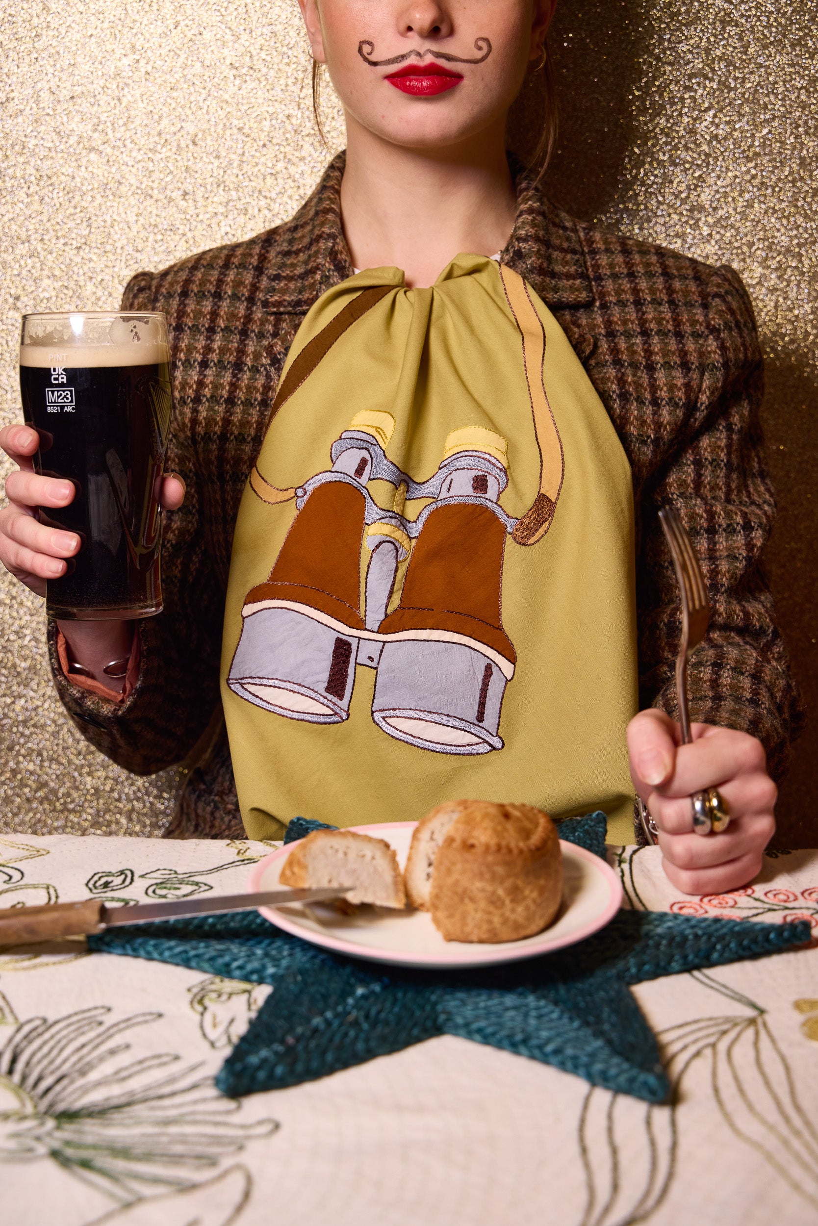 A person wearing a brown tartan jacket and a yellow-green napkin with a binocular graphic, holding a glass of guinness and a fork at a table with bread and a star-shaped placemat. The Jacksons