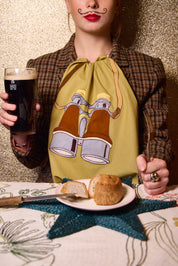 A person wearing a brown tartan jacket and a yellow-green napkin with a binocular graphic, holding a glass of guinness and a fork at a table with bread and a star-shaped placemat. The Jacksons