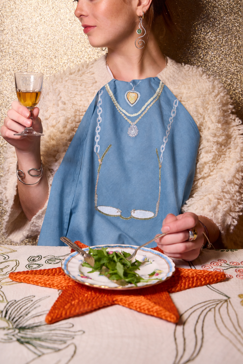 A woman wearing a light blue embroidered napkin with gold and white chain motifs, seated at a table with a plate of salad and a star-shaped orange placemat. The Jacksons London