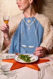 A woman wearing a light blue embroidered napkin with gold and white chain motifs, seated at a table with a plate of salad and a star-shaped orange placemat. The Jacksons London