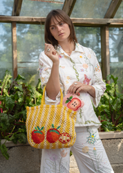 A woman stands in a greenhouse wearing a white floral pajama set and holding a yellow and red jute tote bag with red tomato motif, alongside a matching apple-beaded pouch