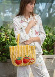 A woman wearing a white floral pajama set holds a yellow jute tote bag decorated with embroidered red apples and a butterfly. She is standing in a greenhouse with green plants in the background.