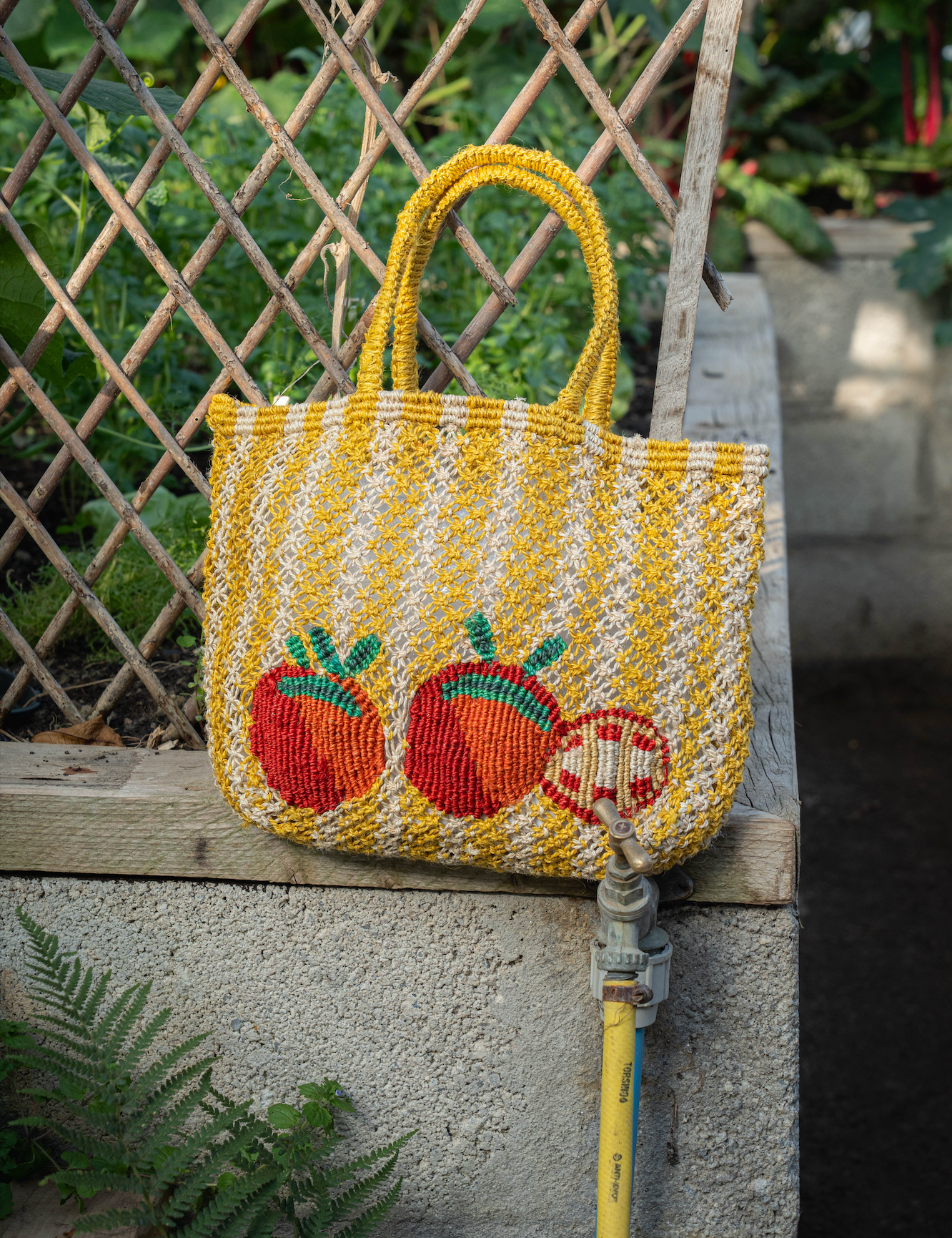 A yellow and white natural striped jute tote bag with embroidered red tomatos and green leaves, resting on a concrete ledge in an outdoor garden setting.