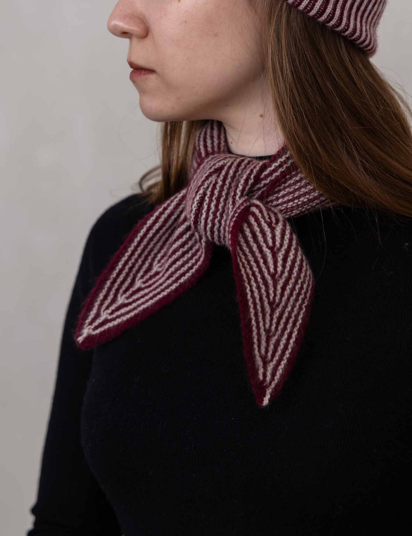 A close-up side profile of a woman wearing a dark knit top and a burgundy and cream striped knitted scarf tied around her neck, with a matching striped beanie partially visible.