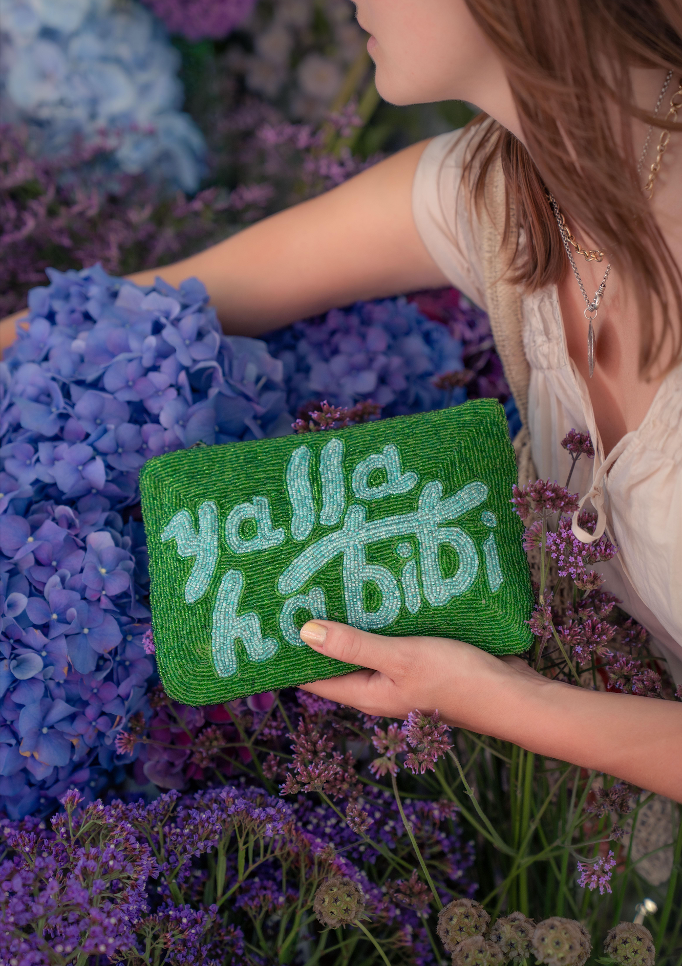 A woman in a cream lace top holds a green beaded clutch bag with text reading 'yalla habibi' amidst purple and blue hydrangea flowers.
