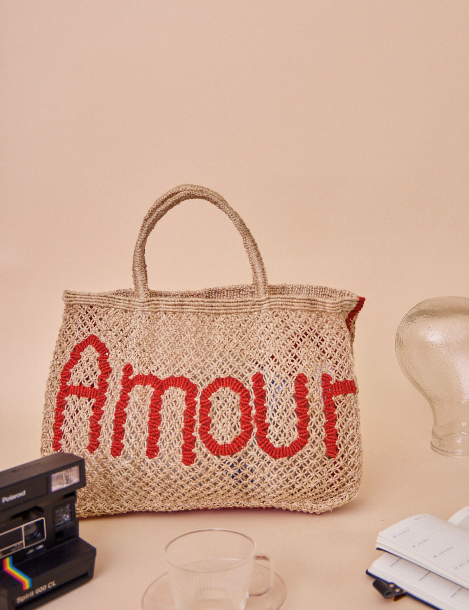 A woven jute bag with the word 'Amour' in red across the front, styled on a beige surface with vintage props including a Polaroid camera, a glass cup, and a notebook.