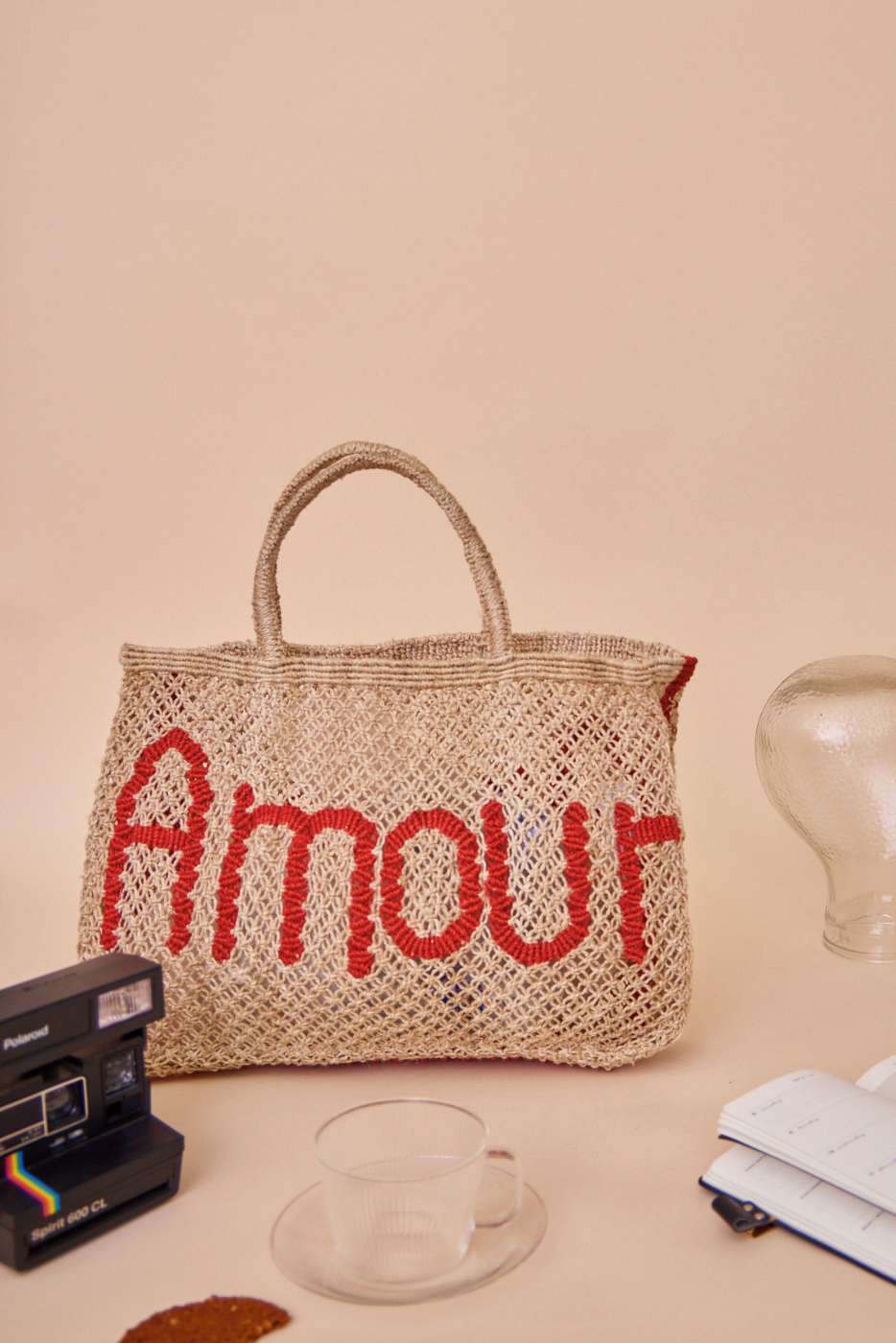 A woven jute bag with the word 'Amour' in red across the front, styled on a beige surface with vintage props including a Polaroid camera, a glass cup, and a notebook.