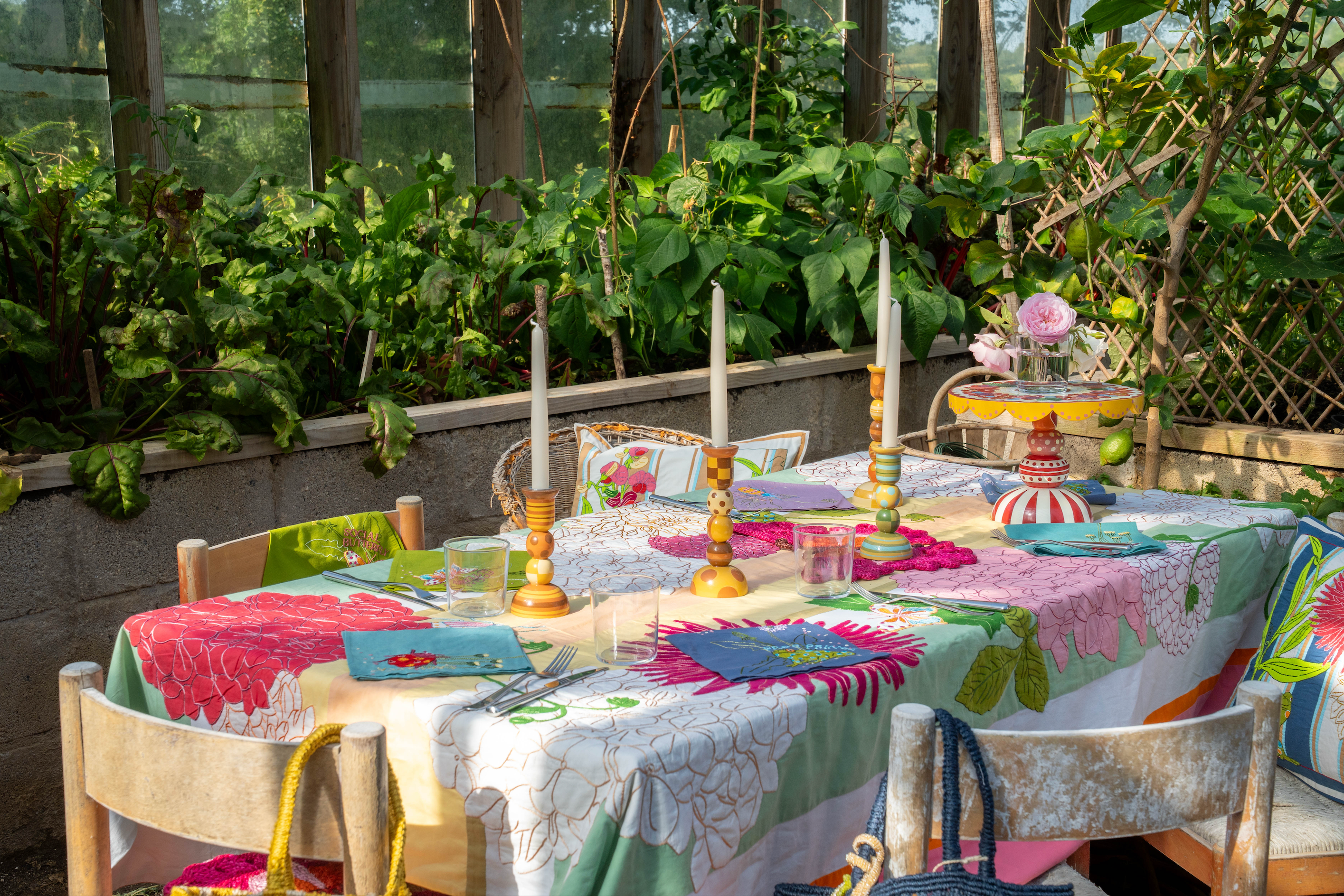A colorful outdoor botanical dining table set in a lush garden greenhouse, featuring a vibrant floral tablecloth, matching chairs, gold candlesticks and decorative objects.
