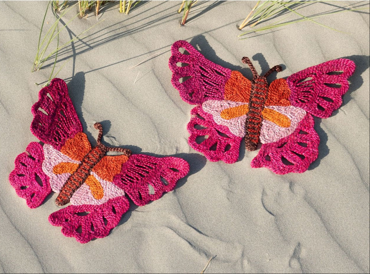 Two pink and orange butterfly-shaped jute placemats on a sandy beach.