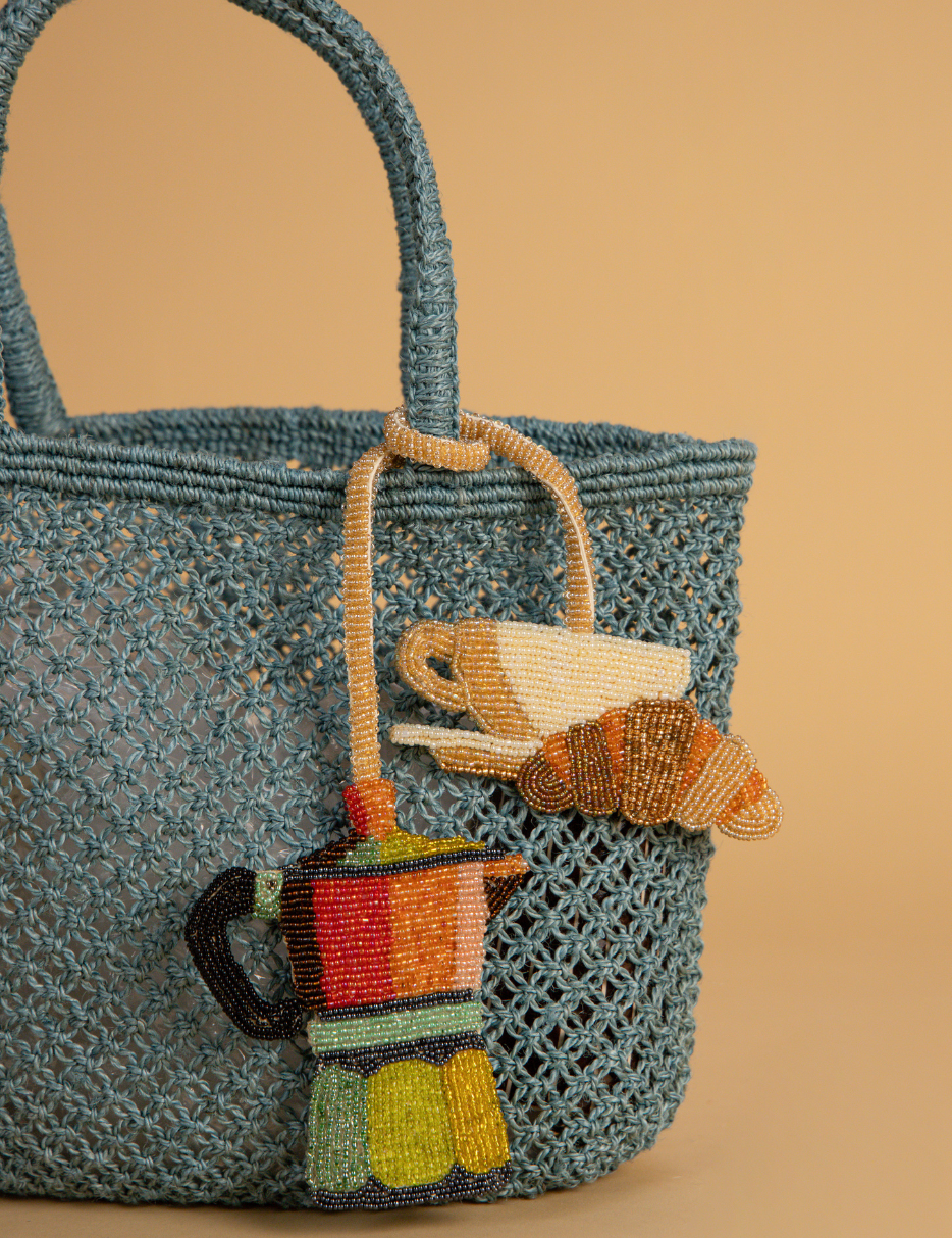 A blue crocheted tote bag with woven handles, decorated with colorful beaded coffee-themed charms including a moka pot, teacup, and croissant, set against a solid beige background.