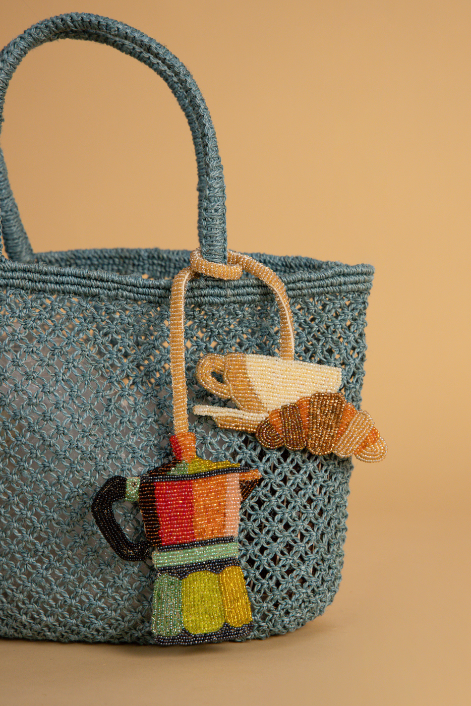 A blue crocheted tote bag with woven handles, decorated with colorful beaded coffee-themed charms including a moka pot, teacup, and croissant, set against a solid beige background.