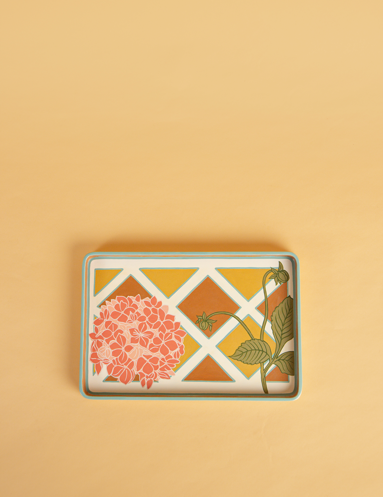 A rectangular decorative tray with a floral and geometric pattern, featuring a pink hydrangea, green leaves, and a white lattice design over a warm yellow and orange background, set against a solid beige backdrop.