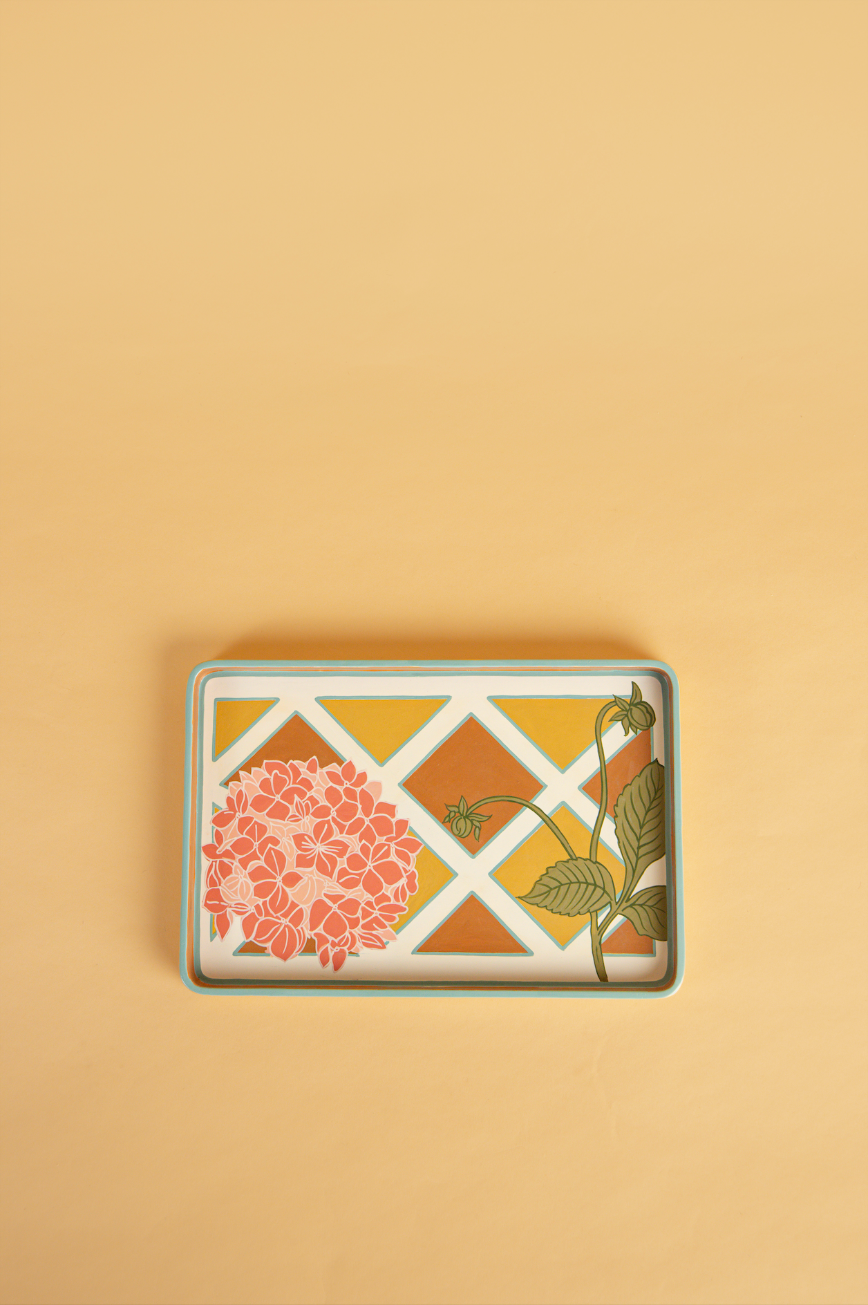 A rectangular decorative tray with a floral and geometric pattern, featuring a pink hydrangea, green leaves, and a white lattice design over a warm yellow and orange background, set against a solid beige backdrop.