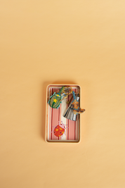 A small handpainted rectangular tray, with a pink and white striped pattern, containing several photographed from above on a solid beige background.