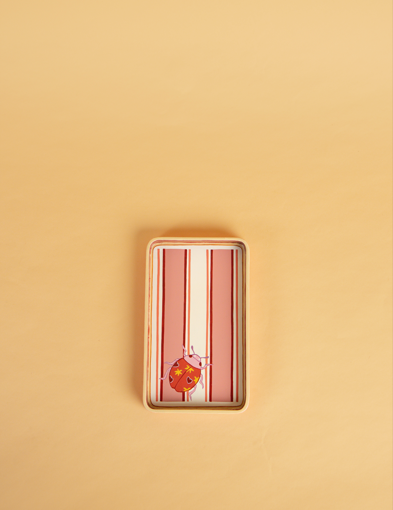A rectangular wooden hand-painted tray with rounded corners, featuring vertical pink and white stripes and a small orange ladybird illustration near the bottom, set against a solid beige background.