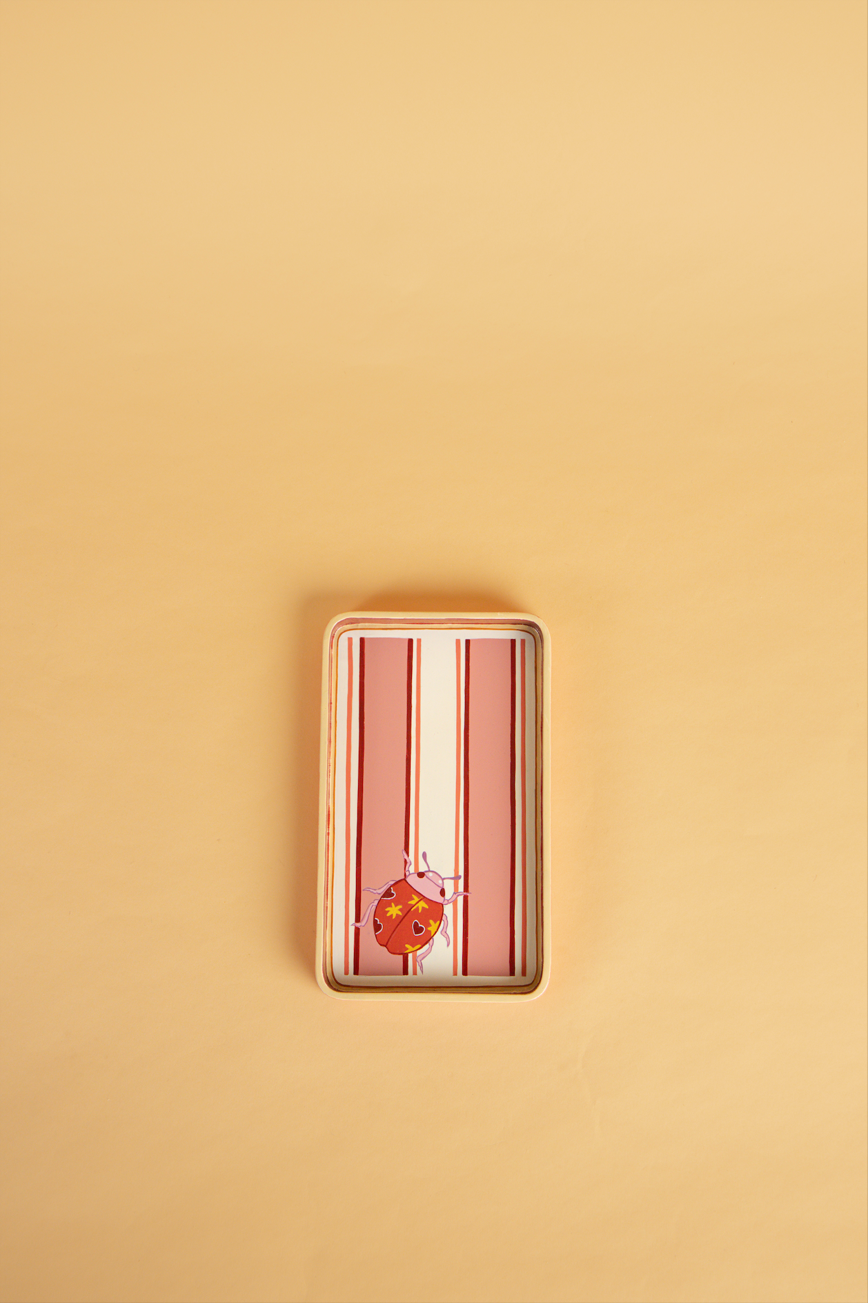 A rectangular wooden hand-painted tray with rounded corners, featuring vertical pink and white stripes and a small orange ladybird illustration near the bottom, set against a solid beige background.