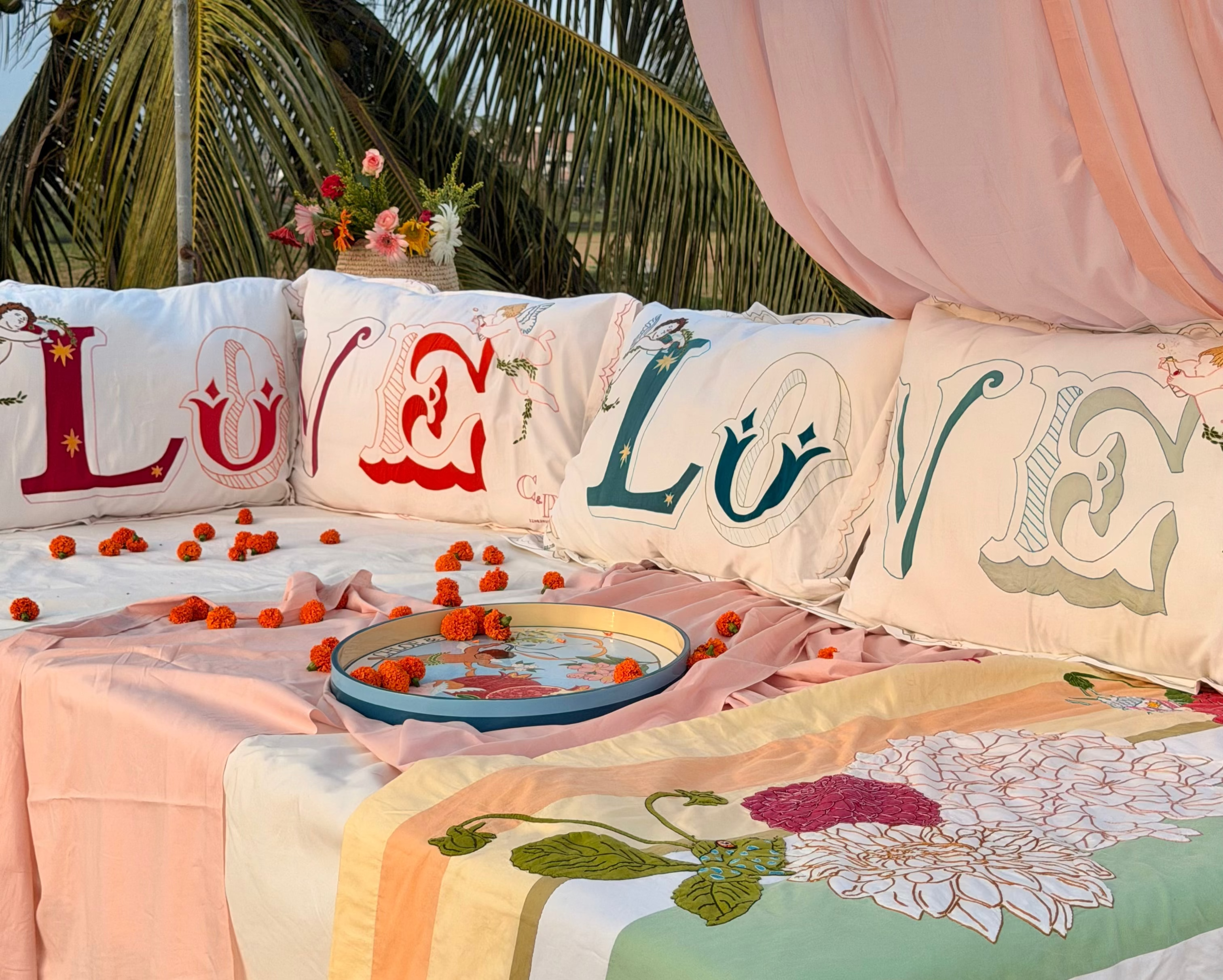 A styled outdoor bed setup draped with pink fabric canopy, decorated with The Jacksons embroidered pillows spelling LOVE, pink sheets, a floral quilt, scattered rose petals, and a wooden Wedding tray in the centre.