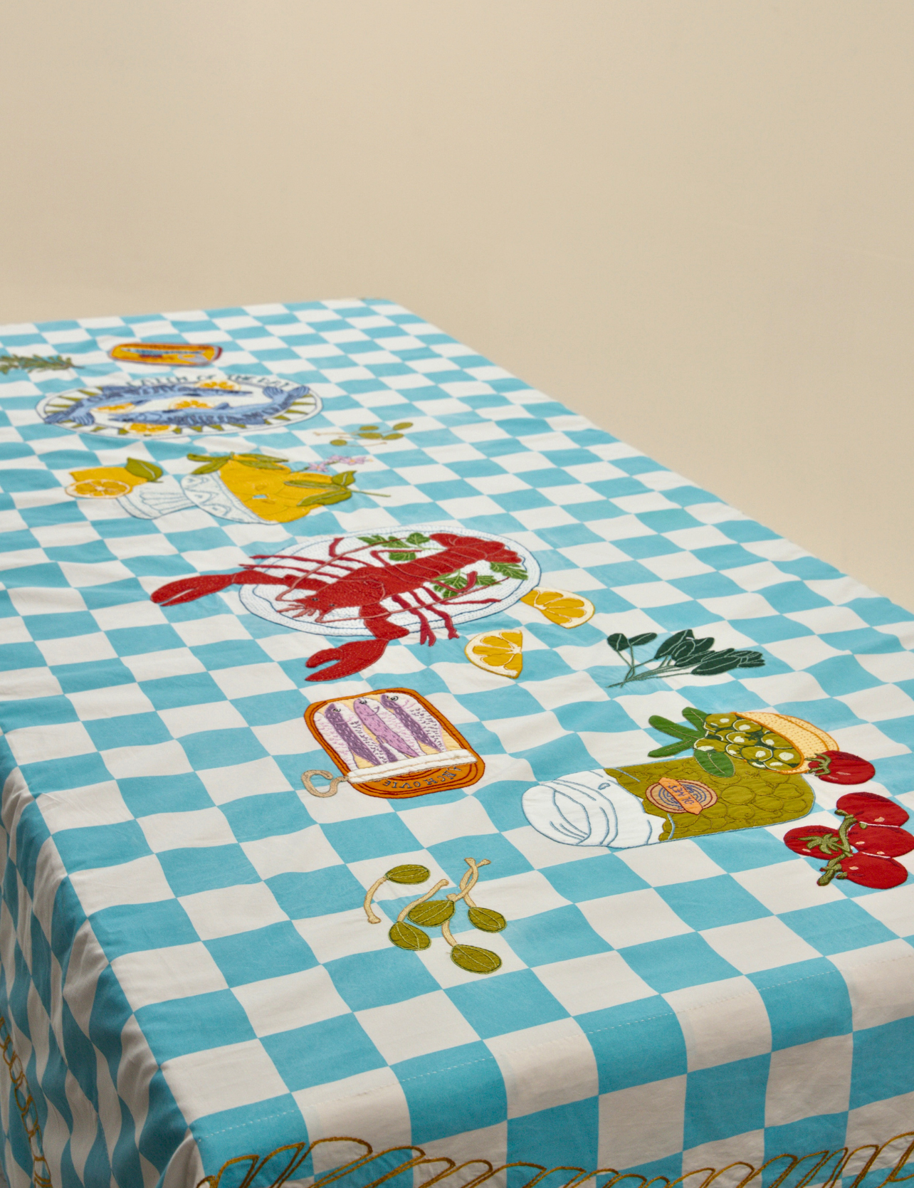 A rectangular table covered with a light blue and white checkered tablecloth featuring embroidered seafood motifs including a red lobster, lemon slices, olives, tomatoes, and a plate of pasta.