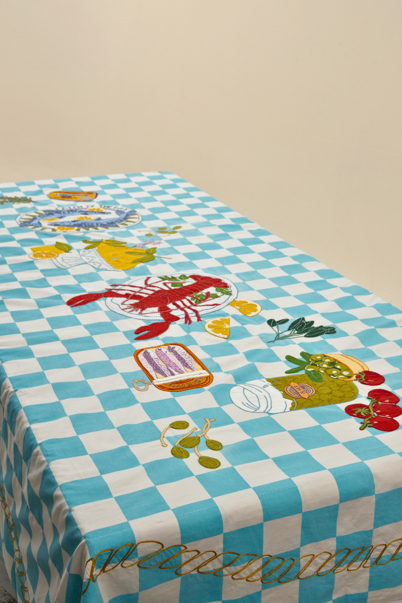 A rectangular table covered with a light blue and white checkered tablecloth featuring embroidered seafood motifs including a red lobster, lemon slices, olives, tomatoes, and a plate of pasta.