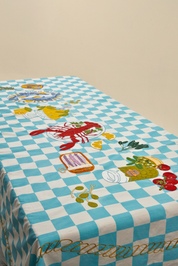 A rectangular table covered with a light blue and white checkered tablecloth featuring embroidered seafood motifs including a red lobster, lemon slices, olives, tomatoes, and a plate of pasta.