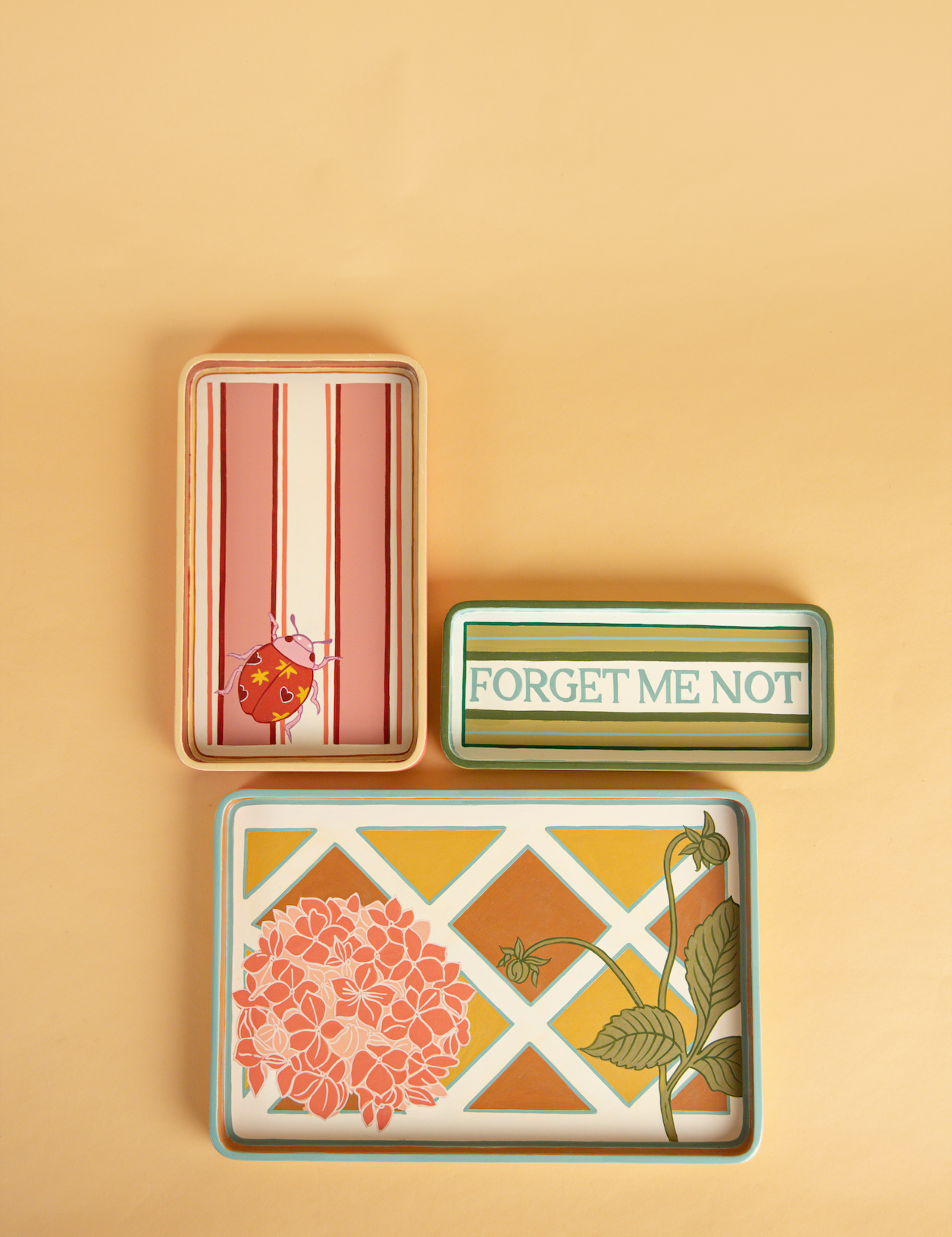 Three hand painted trays. A green blue and white striped tray with forget me not written in elegant lettering. A pink red and white striped tray with a ladybird motif in one corner. A blue, yellow and orange tray with a lattice pattern painted, with a pale pink hydrangea motif in one corner. 