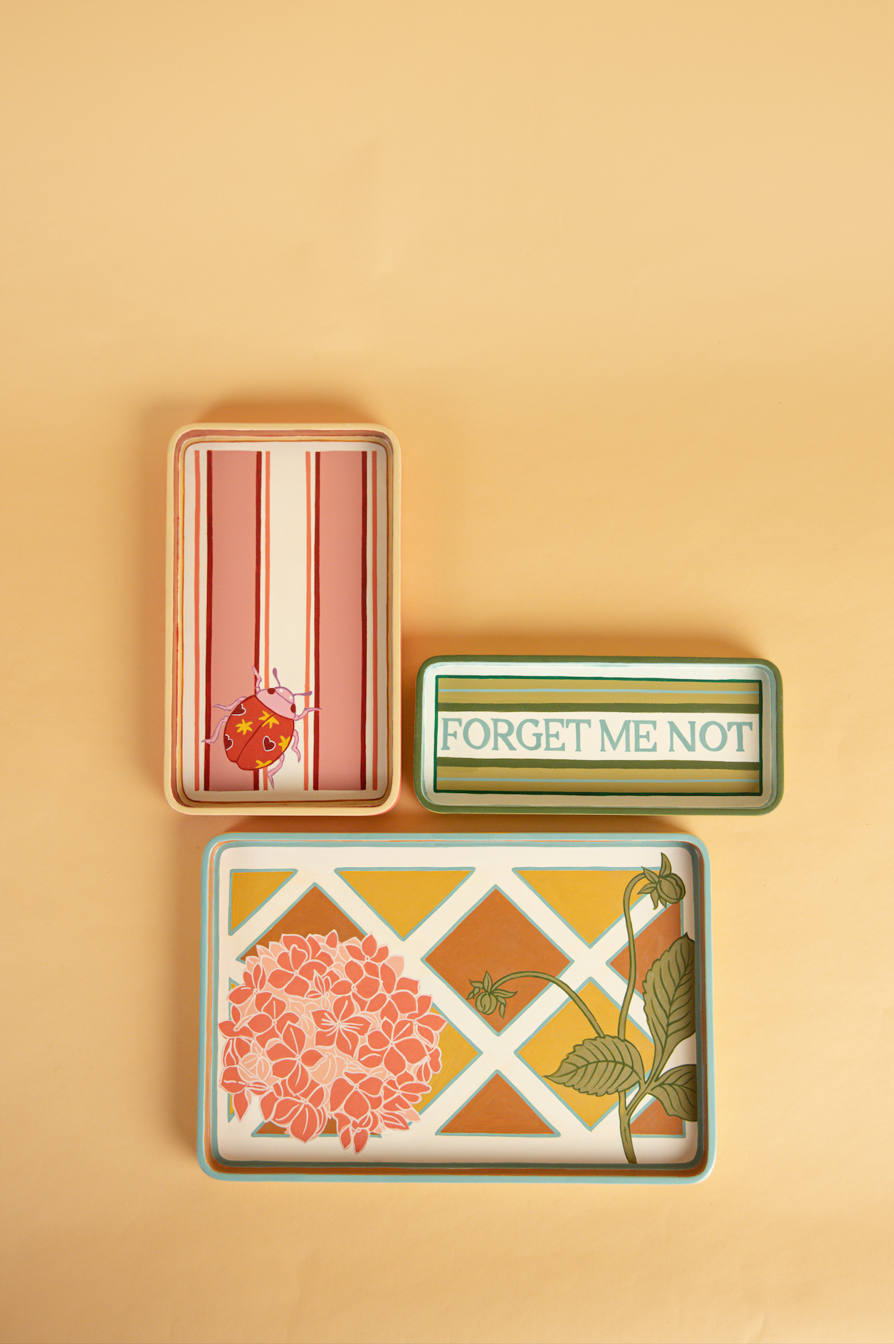 Three hand painted trays. A green blue and white striped tray with forget me not written in elegant lettering. A pink red and white striped tray with a ladybird motif in one corner. A blue, yellow and orange tray with a lattice pattern painted, with a pale pink hydrangea motif in one corner. 